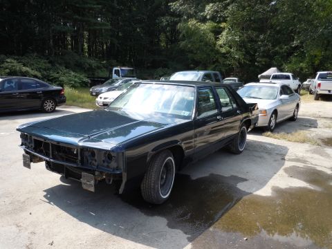 2001 Ford Crown Victoria - A dark sedan, likely a 1991 Ford LTD, sits without a front bumper and grille in an outdoor lot.
