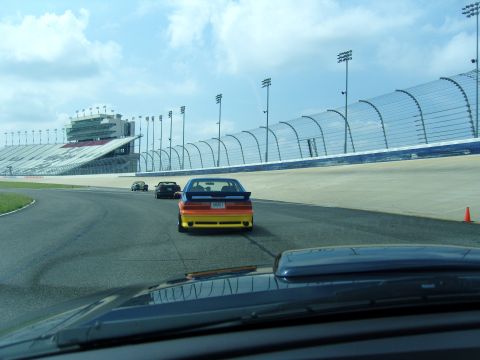 2007 Ford Mustang - Multiple cars race on a track with grandstands under a cloudy sky. The lead car is painted in a rainbow gradient.