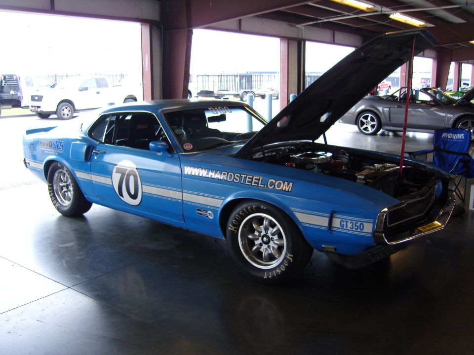 blue 1969 Ford Mustang GT350 race car with hood open, www.hardsteel.com on door, number 70 on side. Another car visible in background