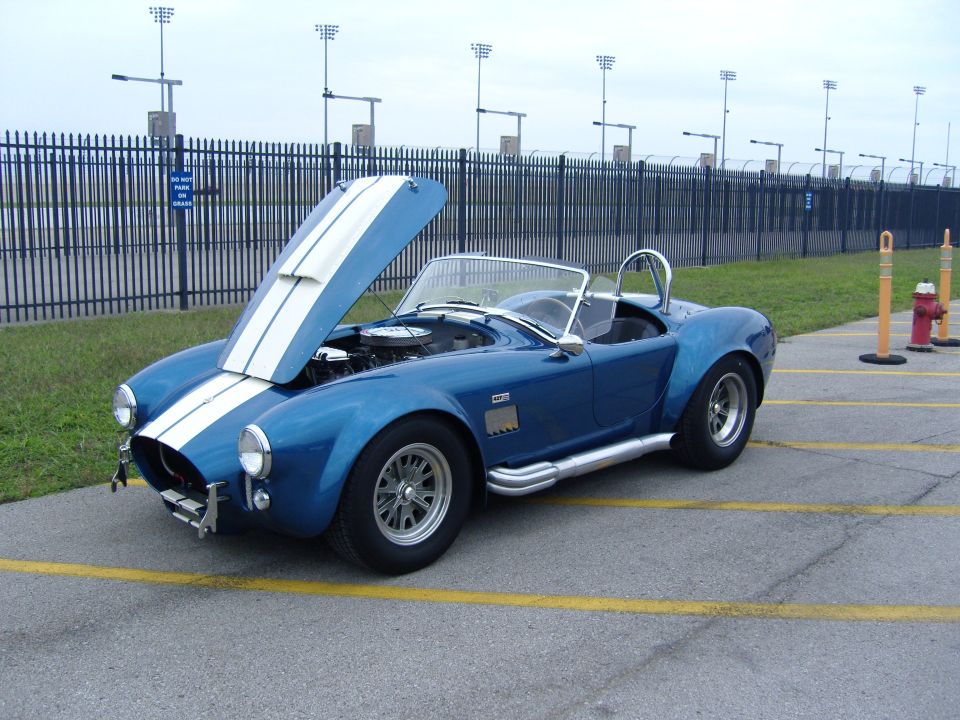 Blue convertible with white racing stripes and hood open, parked on asphalt with yellow lines