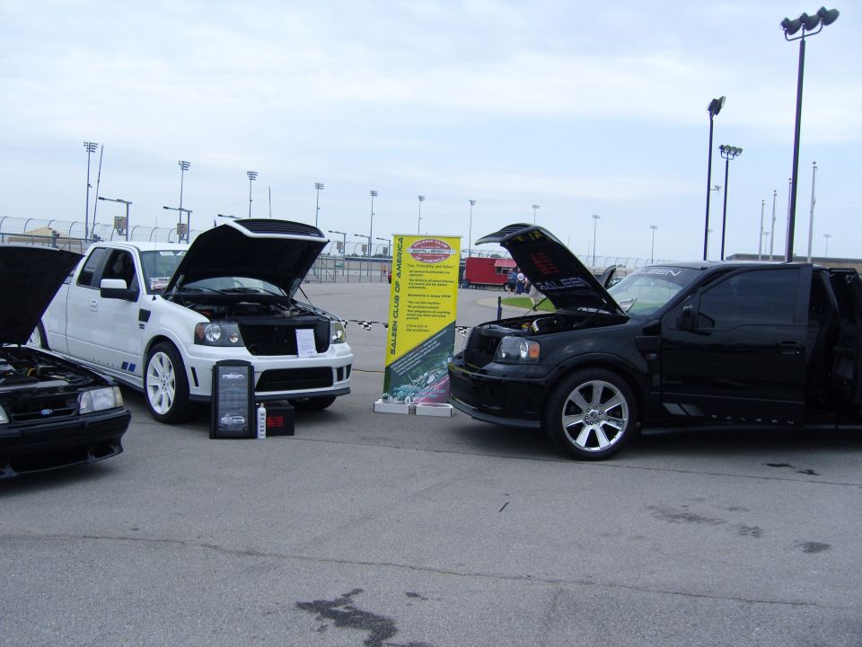 Black and white Ford F-150 trucks with hoods open, plus a black Ford Mustang in foreground. Yellow banner behind trucks