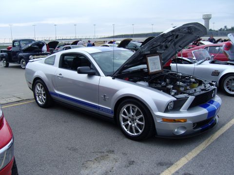 2007 Ford Mustang - A silver 2007 Ford Mustang GT500 with blue racing stripes is displayed with its hood open at a car show, alongside other vehicles.
