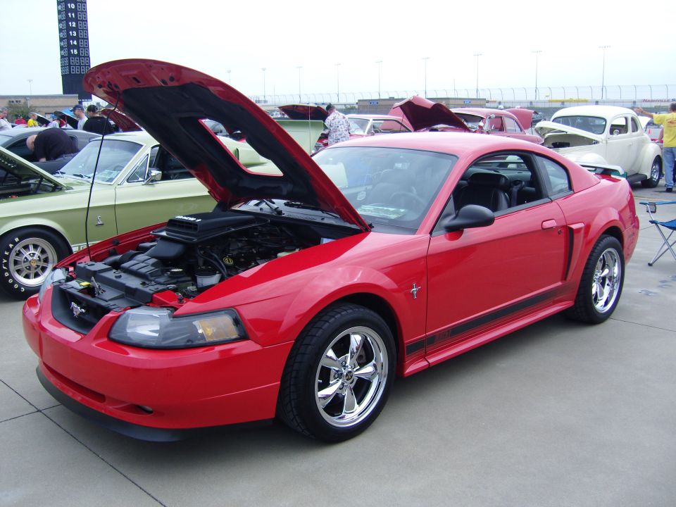 Red Ford Mustang with hood open, showing engine. Another green Mustang is visible to the left
