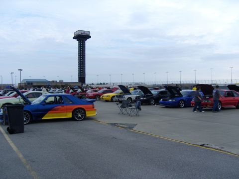 2007 Ford Mustang - A gathering of Mustangs at a car show, with a prominent scoreboard tower overlooking the scene.