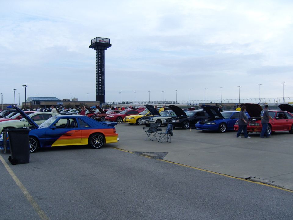 Cars parked in a lot with a tall scoreboard tower in the background. Some hoods are up