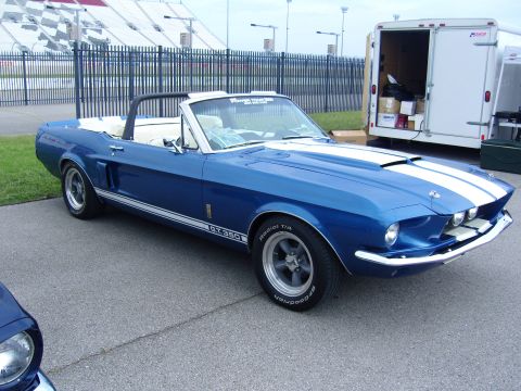 2007 Ford Mustang - A blue convertible Mustang with white racing stripes and GT.350 badging is parked in front of a trailer and racetrack bleachers.