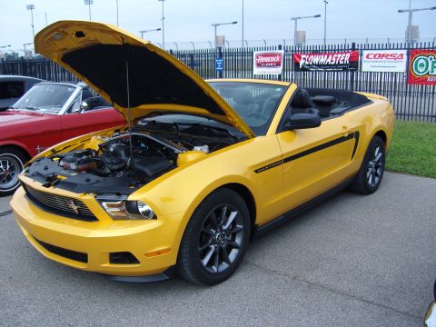 2007 Ford Mustang - Yellow convertible Mustang with black racing stripes and hood open, showing engine. A red classic car is partially visible to the left.