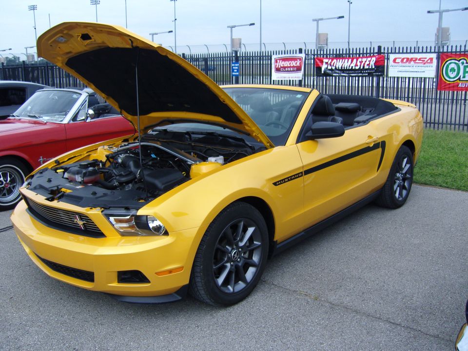 Yellow convertible Mustang with black racing stripes and hood open, showing engine. A red classic car is partially visible to the left