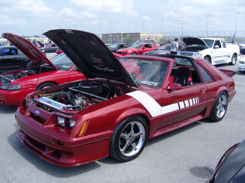 2007 Ford Mustang - A red 1985 Ford Mustang GT coupe with white racing stripes is displayed with its hood open at a car show.