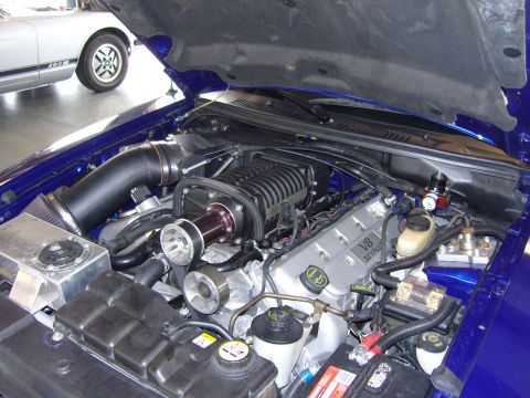 2007 Ford Mustang - The engine bay of a blue vehicle, a prominent supercharger and V8 32V engine components. A second vehicle is partially visible in the backg…