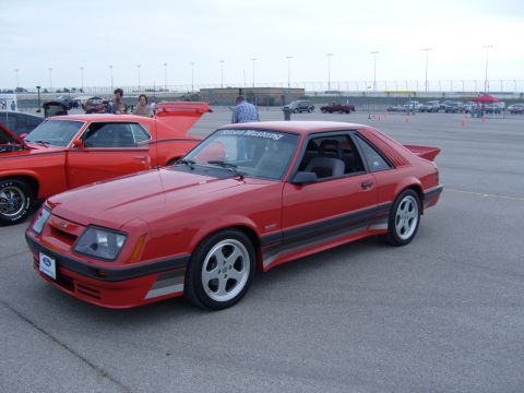2007 Ford Mustang - A red coupe car with a spoiler and racing stripes is parked on asphalt, with other vehicles in the background.