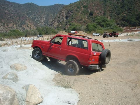 1988 Ford Bronco - A red SUV navigates a rocky trail with mountains in the distance and other vehicles present.