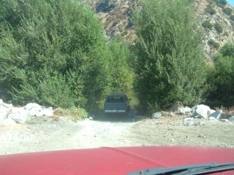 1988 Ford Bronco - SUV navigates a dirt path and shallow stream flanked by dense trees and rocky hills.