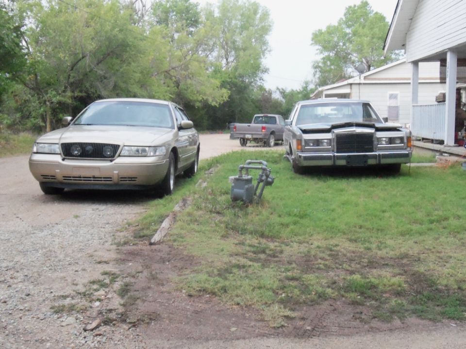Gold sedan with quad headlights parked on gravel, silver sedan with hood up on grass, pickup truck in background