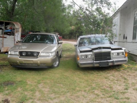 2000 Mercury Grand Marquis - Two sedans are parked on a grassy area next to a building, with trees in the background.