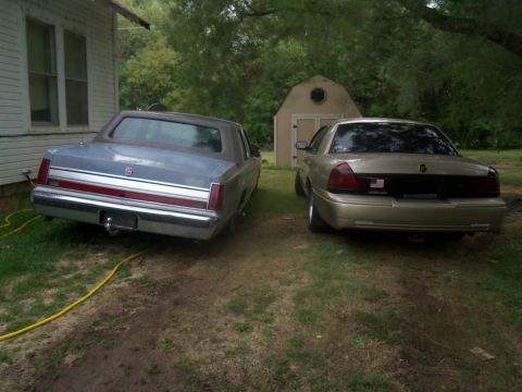 2000 Mercury Grand Marquis - Two sedans are parked in a yard with a house and shed in the background.