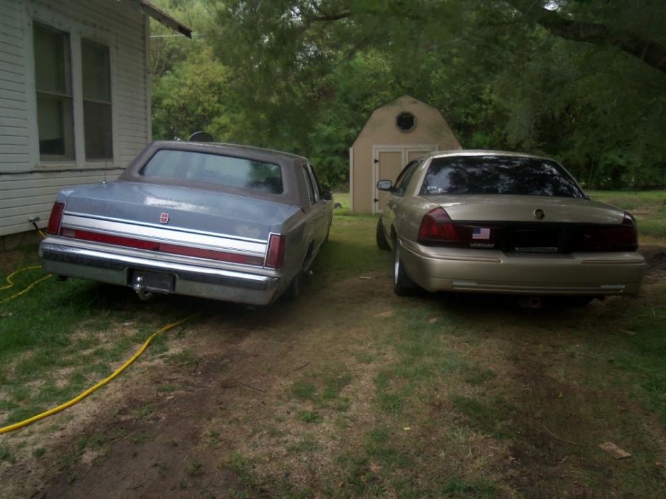 Gold sedan and a blue sedan parked in a grassy yard next to a house and shed