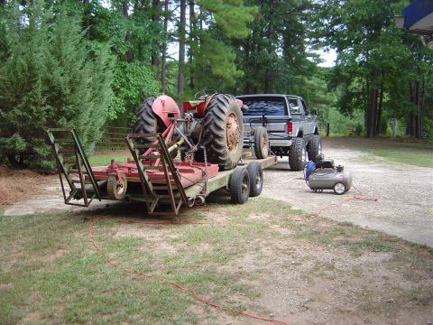 1986 Ford Bronco - A silver pickup truck is seen towing a trailer loaded with a tractor, with an air compressor nearby.