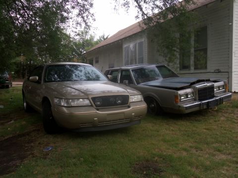 2000 Mercury Grand Marquis - Two sedans are parked on a grassy area next to a house. The front vehicle is gold, the rear is gray.