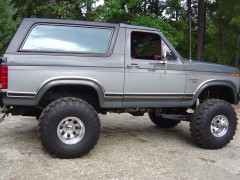 1986 Ford Bronco - This lifted 1986 Ford Bronco is shown from the side, parked outdoors on a gravelly surface with trees in the background.