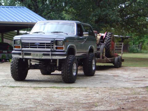 1986 Ford Bronco - A lifted 1986 Ford Bronco is shown towing a tractor on a trailer, likely for a farm or off-road excursion.