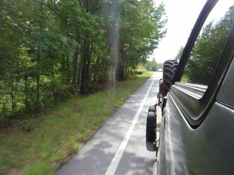 1986 Ford Bronco - Two vehicles travel down a two-lane road bordered by trees, with one vehicle's spare tire visible.