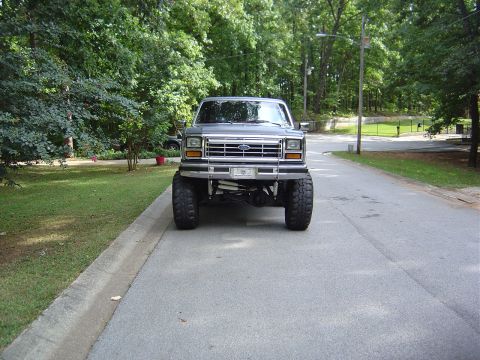1986 Ford Bronco - This lifted Ford truck is shown from the front on a paved road with trees and grass in the background.