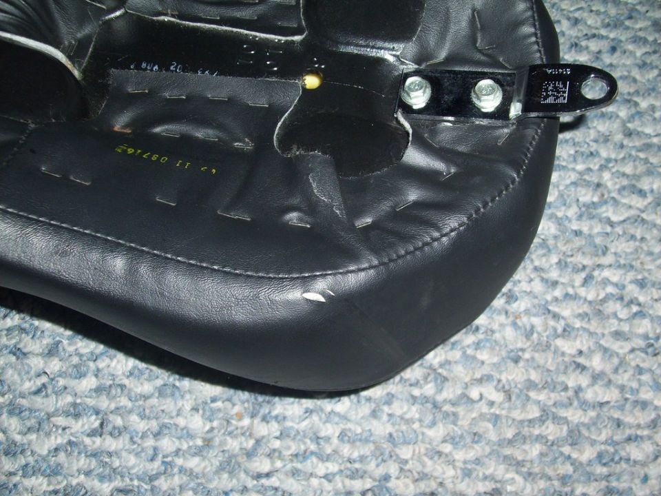 Underside of a black motorcycle seat showing seat pan staples and bolted mounting tab
