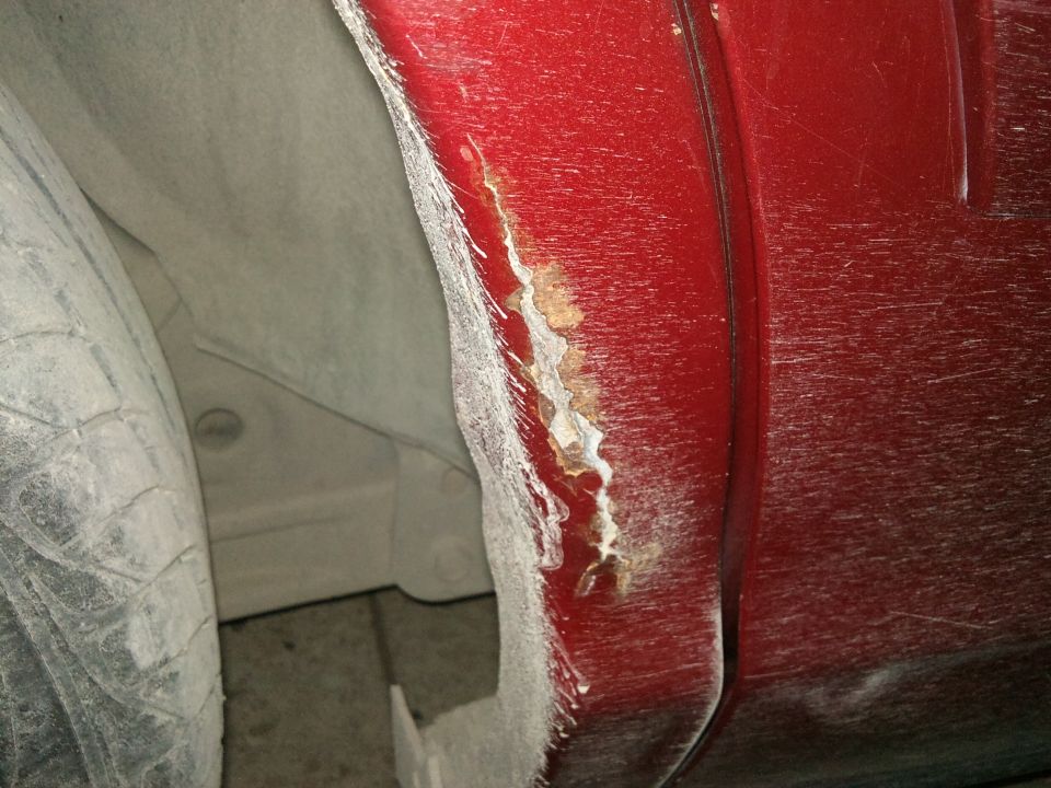 Close-up of rust on a red car's rear wheel well. Tire is visible on the left