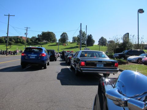 1991 Ford LTD Crown Victoria - black Highway Patrol sedan waits in a line of vehicles on a road with a green hillside and parked motorcycles behind.