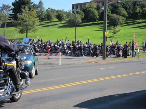 1991 Ford LTD Crown Victoria - motorcycle rider in the foreground faces a crowd gathered with parked motorcycles along a grassy hillside by a road.