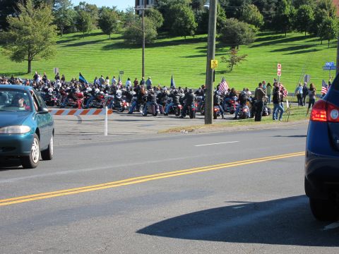 1991 Ford LTD Crown Victoria - Cars travel a road past a gathering of people and parked motorcycles flying flags on a grassy hillside.