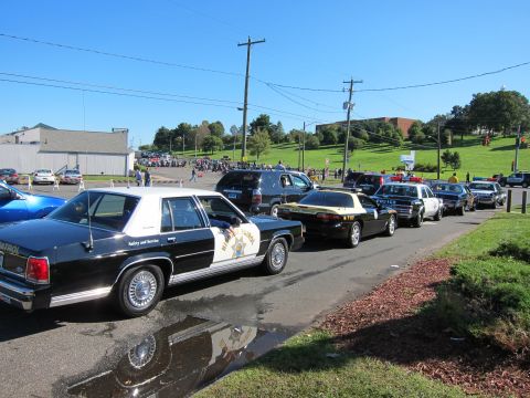 1991 Ford LTD Crown Victoria - black-and-white Highway Patrol sedan leads a line of classic police cars parked along a lot, with a crowd on a hill behind.