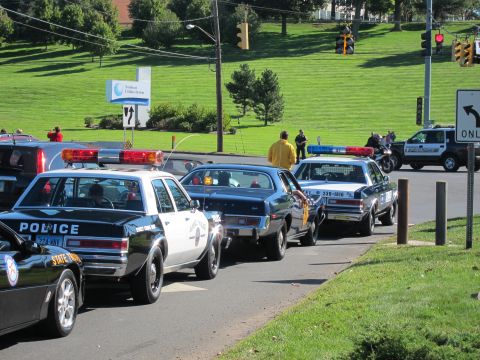 1991 Ford LTD Crown Victoria - black-and-white vintage Police sedan and a maroon coupe sit in a curving line of classic patrol cars beside a grassy slope.