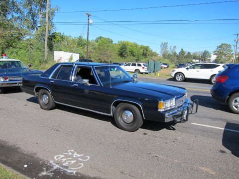 1991 Ford LTD Crown Victoria - dark blue vintage Ford sedan fitted with a front push bar drives along a road past parked cars and roadside greenery.
