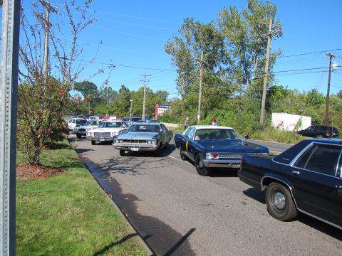 1991 Ford LTD Crown Victoria - line of vintage police cars, including dark sedans with light bars, stretches along a tree-lined road under blue sky.