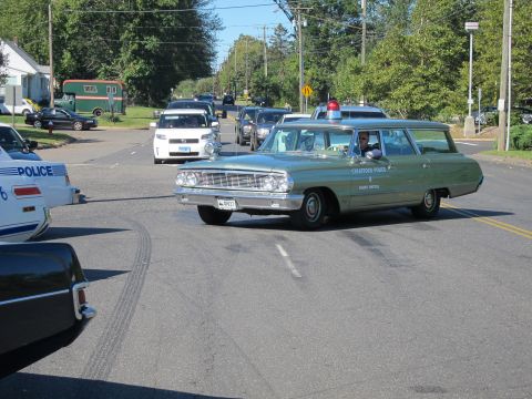 1991 Ford LTD Crown Victoria - green 1960s Ford station wagon lettered Stratford Police turns through an intersection past other parked classic cars.
