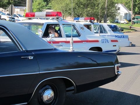 1991 Ford LTD Crown Victoria - White vintage police cars with red roof light bars line up, one numbered 1776, beside a dark sedan in the foreground.