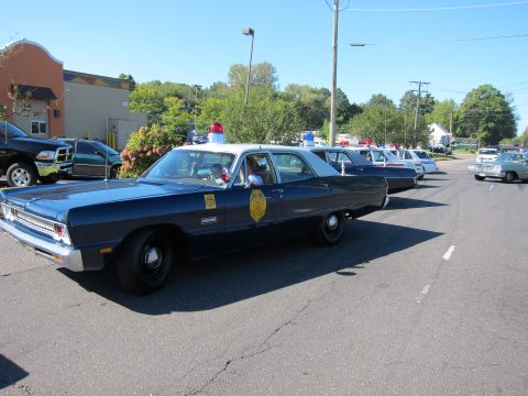 1991 Ford LTD Crown Victoria - dark blue vintage state patrol sedan with a gold door emblem and red roof light heads a line of classic police cars in a lot.