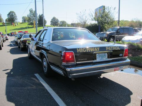 1991 Ford LTD Crown Victoria - Rear view of a black Ford LTD Crown Victoria Highway Patrol cruiser driving on a road ahead of more classic patrol cars.