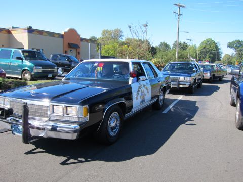 1991 Ford LTD Crown Victoria - black-and-white Ford Crown Victoria Highway Patrol car with push bar leads a line of classic patrol cars across a parking lot.