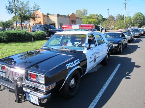 1991 Ford LTD Crown Victoria - black-and-white vintage Police sedan with a front push bar and red roof light bar leads a line of cars in a parking lot.