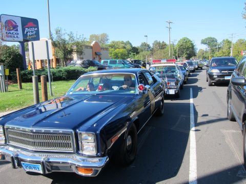 1991 Ford LTD Crown Victoria - dark blue classic police sedan with a person at the wheel leads a procession of vehicles through a parking lot past fast-food signs.
