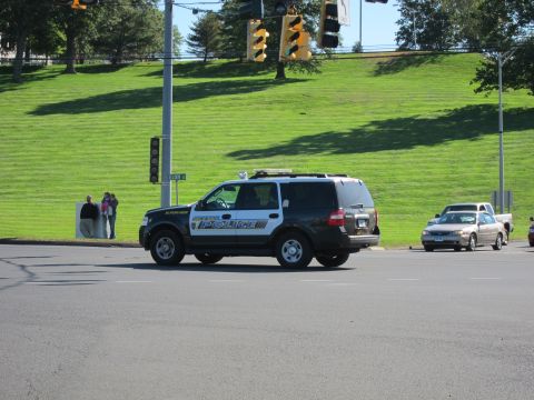 1991 Ford LTD Crown Victoria - black and white POLICE SUV crosses a broad intersection with traffic signals, a grassy slope and pedestrians in the background.