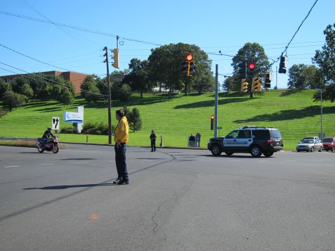 1991 Ford LTD Crown Victoria - man in a yellow shirt stands in a wide intersection directing traffic as a motorcycle and an SUV approach under red signals.