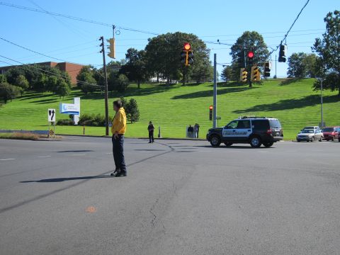 1991 Ford LTD Crown Victoria - man in a yellow shirt directs traffic at an intersection while a police SUV and other cars wait near a grassy hillside.
