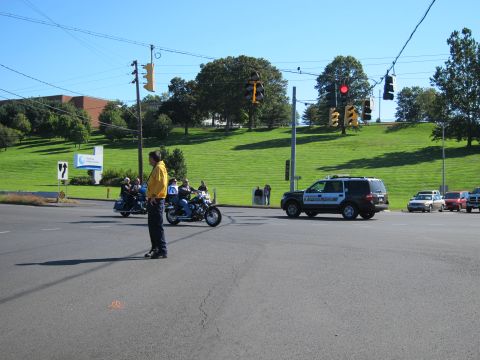 1991 Ford LTD Crown Victoria - man in a yellow shirt directs a group of motorcycles and a police SUV across an intersection beside a green hill.