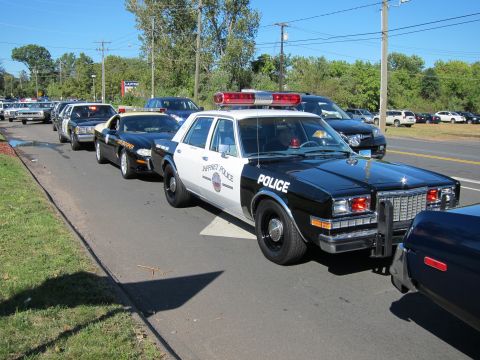 1991 Ford LTD Crown Victoria - black and white police sedan with red dome light heads a line of parked patrol cars along a curb on a sunny day.