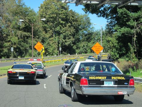 1991 Ford LTD Crown Victoria - black HIGHWAY PATROL sedan and other police cars drive along a road past orange right-lane-closed warning signs.