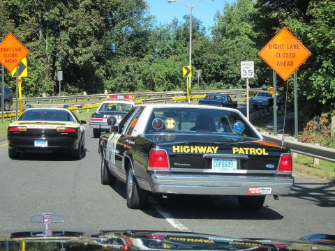 1991 Ford LTD Crown Victoria - View through a windshield of a black HIGHWAY PATROL sedan and other cruisers driving past orange RIGHT LANE CLOSED AHEAD signs.